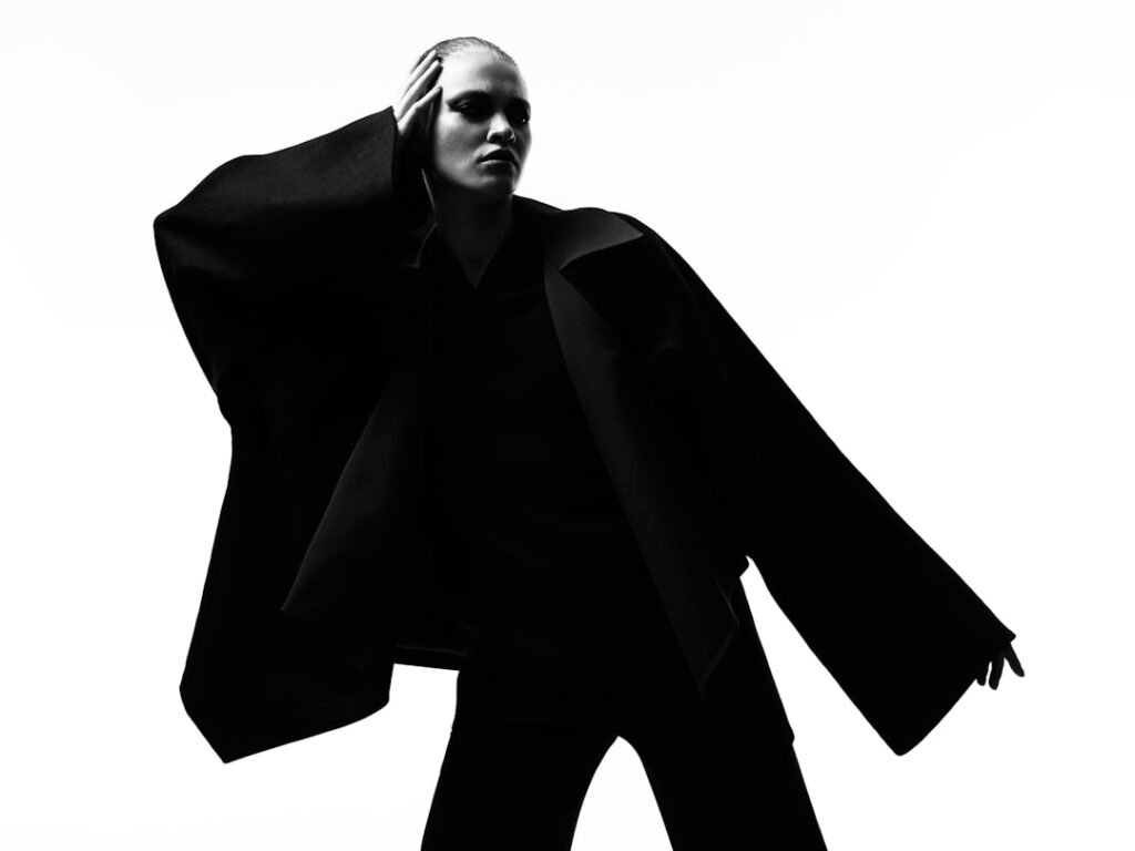 Black and white editorial fashion photograph of a model in a dramatic oversized structured black coat, representing the Antwerp Six's architectural, concept-driven aesthetic