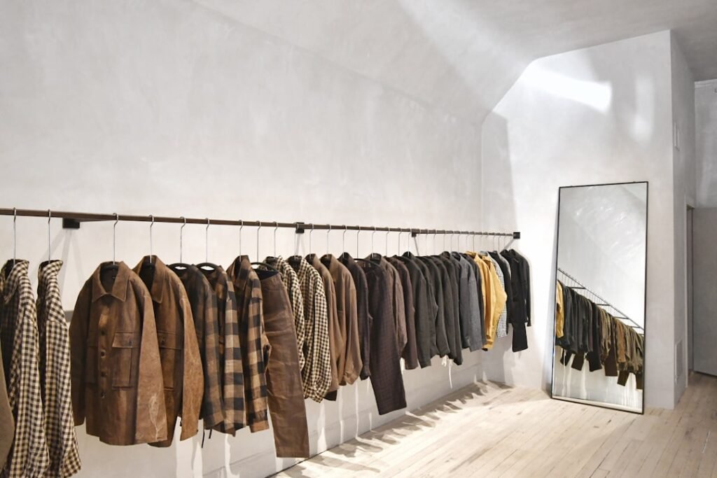 A minimalist luxury fashion atelier interior with a rail of tailored menswear jackets in muted earth tones against a whitewashed wall, evoking the pared-back aesthetic of Antwerp designer spaces