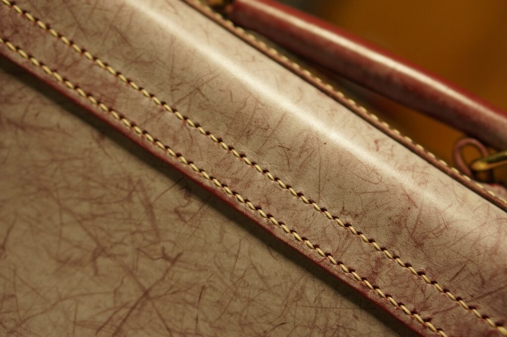 Extreme close-up of aged vachetta-style leather on a luxury bag showing the natural patina, yellowed tone, and fine stitching along the seam