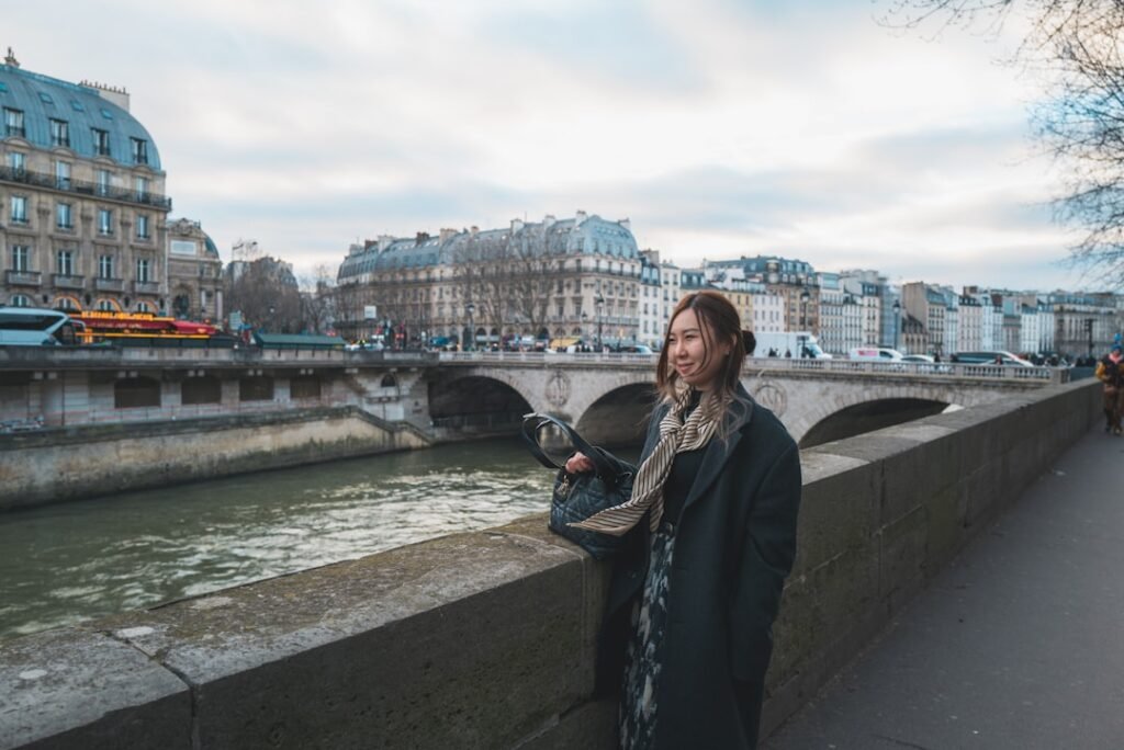 Woman in a long dark wool coat and scarf standing by the Seine river in Paris in winter, with Haussmann buildings in the background.